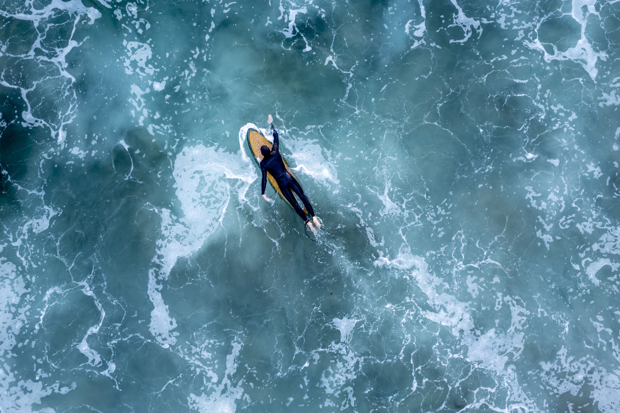 Aerial view of surfer paddling on a surfboard in turquoise ocean waves.
