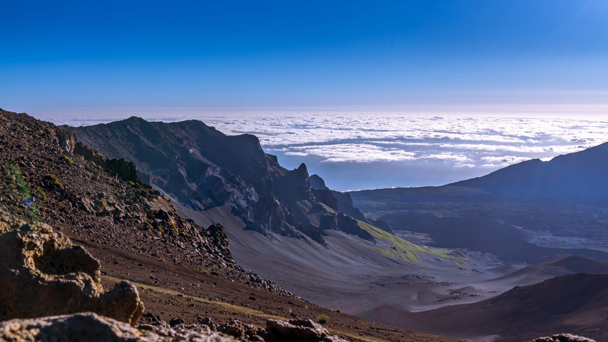 Mountain landscape with rocky cliffs and a sea of clouds under a clear blue sky.