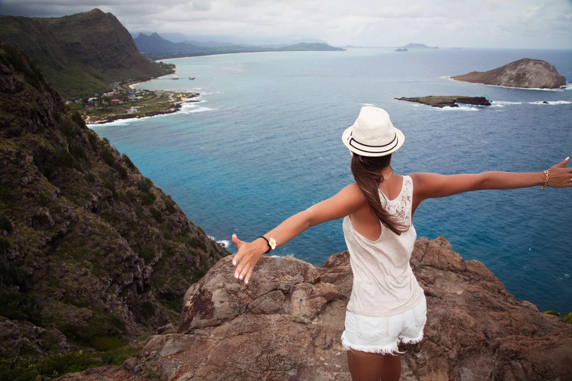 Woman with hat enjoying ocean view from cliff with arms outstretched.