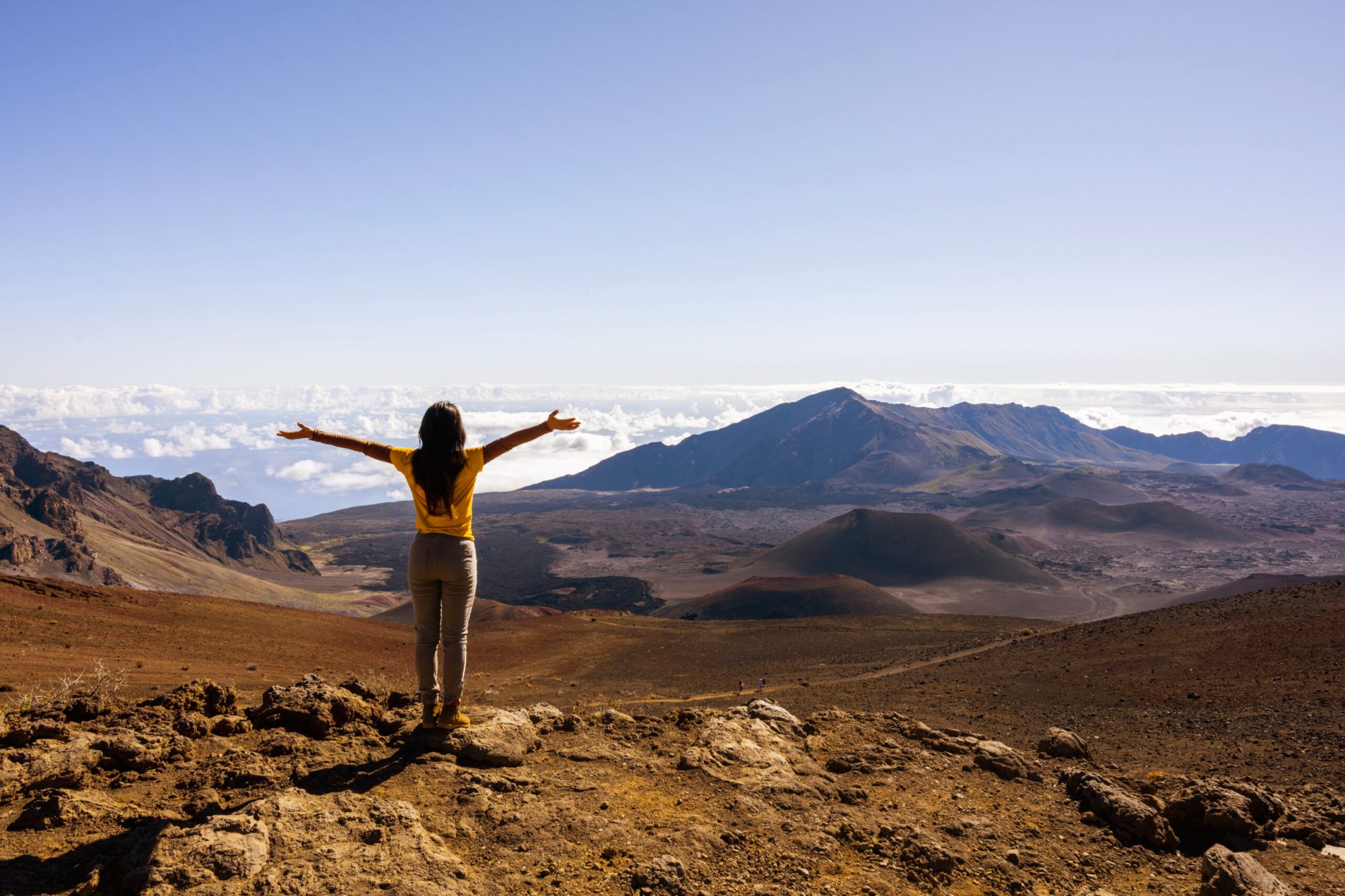 Person in yellow stands on hill overlooking a mountain and clouds.