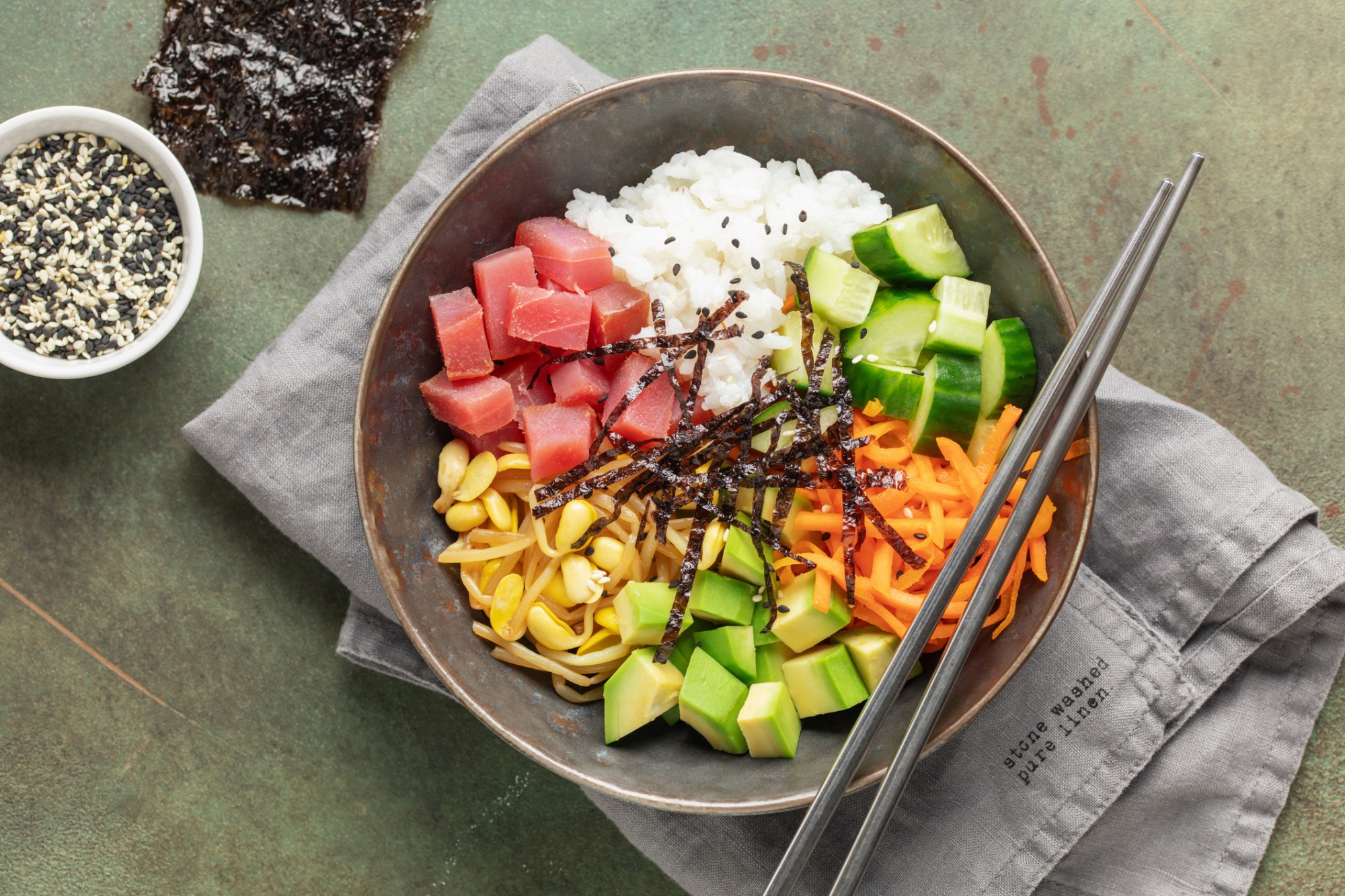 Poke bowl with tuna, rice, cucumber, avocado, carrots, seaweed, and chopsticks on a napkin.