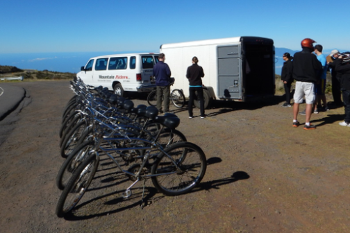 Row of bicycles on roadside with van, trailer, and people in helmets nearby.