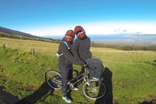 Two people in helmets pose with a bike on a grassy hillside with a scenic view.