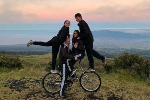 Four people pose on a hilltop; one on a bike, others balanced, with a scenic backdrop of hills and sky.