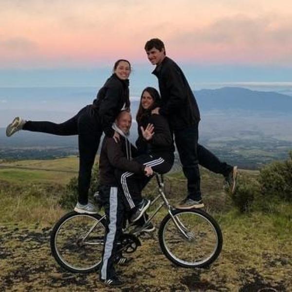 Four people pose on a hilltop; one on a bike, others balanced, with a scenic backdrop of hills and sky.