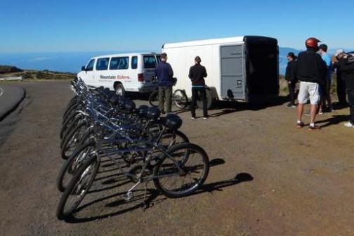 Group of cyclists gather near van and trailer on roadside with row of bikes.